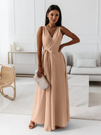 Femme portant une robe longue beige élégante, tenant un sac à main texturé, posant dans un salon moderne. Mode féminine, tenue chic, style contemporain.
