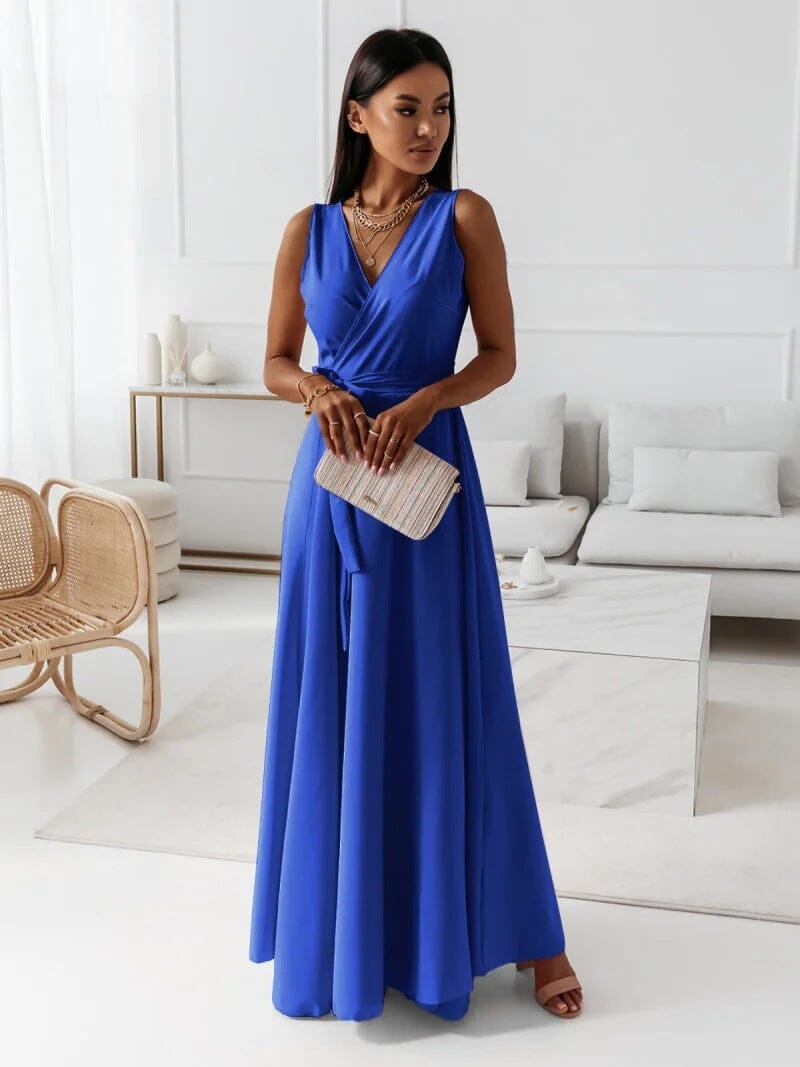Femme en robe longue bleue élégante, tenant une pochette beige, dans un salon moderne avec fauteuil en rotin et canapé blanc. Mode chic et tendance.