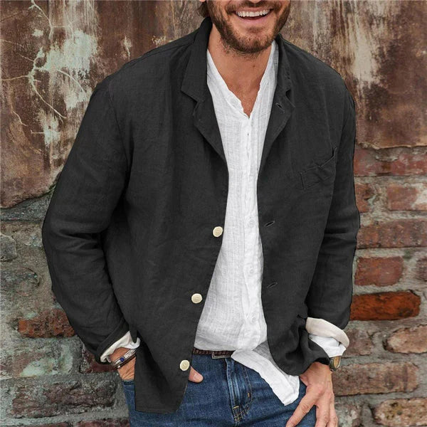 Homme souriant portant une veste noire en lin avec chemise blanche, devant un mur en briques. Mode masculine décontractée, style urbain chic.