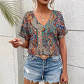 Femme portant un haut à motifs bohèmes colorés et un short en jean déchiré, accessoirisée d'un collier et d'une ceinture, devant un mur texturé.