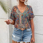 Femme portant un haut à motifs bohèmes colorés et un short en jean déchiré, accessoirisée d'un collier et d'une ceinture, devant un mur texturé.
