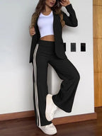 Femme en tailleur noir moderne avec bande blanche, t-shirt blanc, baskets blanches, posant contre un mur intérieur en bois. Mode décontractée élégante.