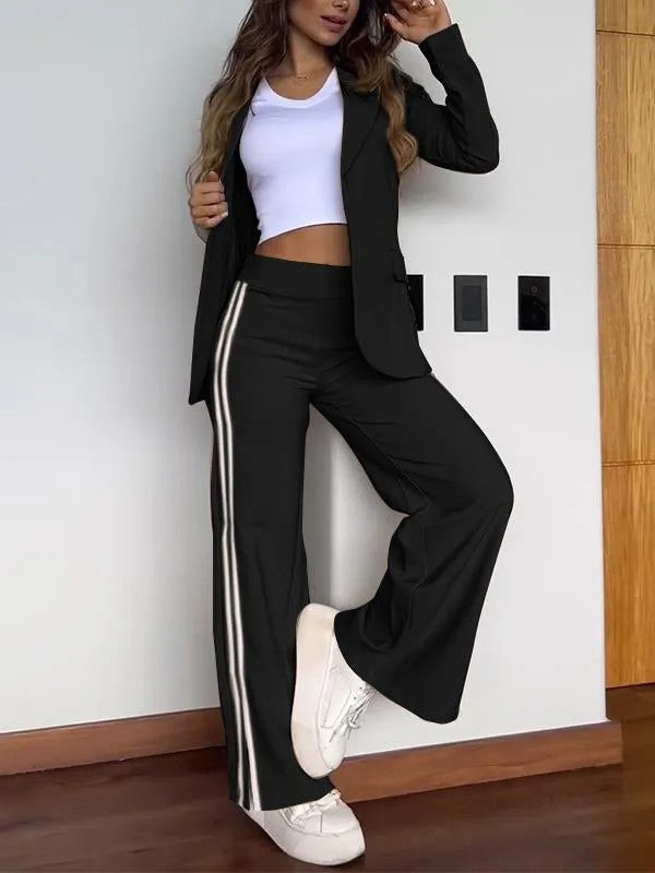 Femme en tailleur noir moderne avec bande blanche, t-shirt blanc, baskets blanches, posant contre un mur intérieur en bois. Mode décontractée élégante.