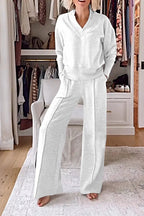 Femme portant un ensemble blanc confortable dans un dressing moderne, avec des vêtements suspendus et une chaise blanche. Mode détente élégante.
