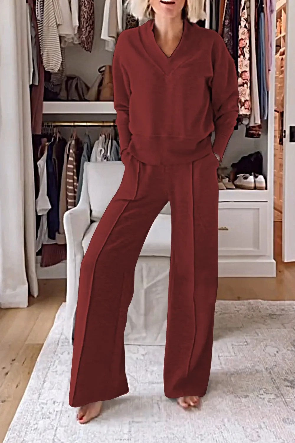 Femme portant un ensemble lounge rouge bordeaux dans un dressing moderne, avec des vêtements suspendus et un fauteuil blanc en arrière-plan.
