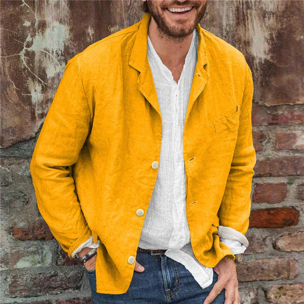 Homme portant une veste jaune en lin, chemise blanche et jeans, posant devant un mur en briques. Mode masculine décontractée, style urbain.