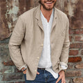 Homme souriant portant une veste en lin beige sur une chemise blanche, posant devant un mur en briques. Mode décontractée, style masculin élégant.