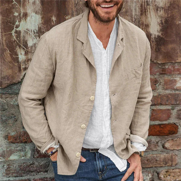 Homme souriant portant une veste en lin beige sur une chemise blanche, posant devant un mur en briques. Mode décontractée, style masculin élégant.