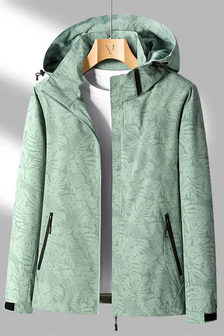 Veste imperméable verte à motifs feuillages, capuche ajustable, fermetures éclair noires, accrochée sur cintre en bois. Mode outdoor tendance.
