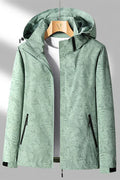 Veste imperméable verte à motifs feuillages, capuche ajustable, fermetures éclair noires, accrochée sur cintre en bois. Mode outdoor tendance.