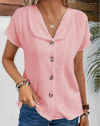 Femme portant une chemise rose en coton gaufré avec col large et boutons en bois, accessoirisée de bracelets, devant un fond végétal.