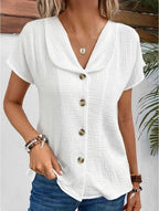 Femme portant un chemisier blanc en coton à boutons, manches courtes, col large, avec un collier doré et des bracelets, style décontracté chic.