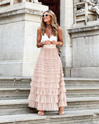Femme souriante en jupe longue à volants rose et haut blanc, portant des lunettes de soleil, posant sur des marches en pierre devant un bâtiment historique.