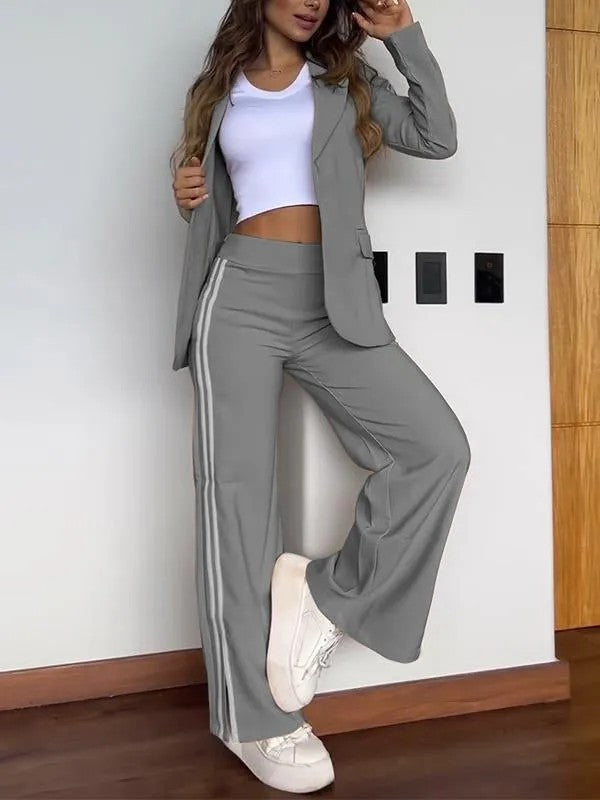 Femme en tailleur gris moderne avec veste et pantalon à rayures, portant un top blanc et des baskets, posant contre un mur intérieur.