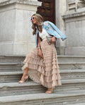 Femme souriante en jupe à volants beige, veste en jean, lunettes de soleil, tenant une pochette, posant sur des marches en pierre devant une porte en bois.