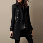 Femme portant un manteau noir élégant à double boutonnage, style trench-coat, sur fond beige. Mode automne-hiver, tendance vestimentaire chic.