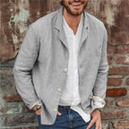 Homme souriant portant une veste en lin gris sur une chemise blanche, posant devant un mur en briques. Mode masculine décontractée, style urbain.
