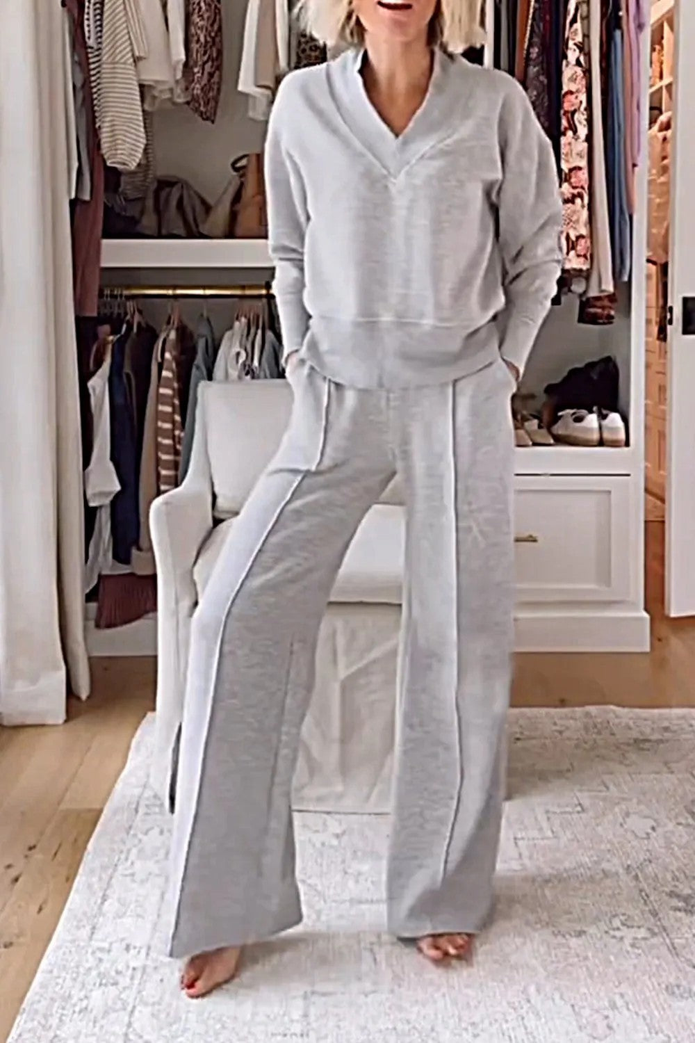 Femme portant un ensemble confortable gris dans un dressing moderne, avec des vêtements suspendus et un fauteuil blanc. Mode détente élégante.