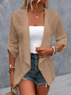 Femme portant un cardigan beige en tricot, débardeur blanc, short en jean, accessoires bracelets, collier doré, style décontracté chic.