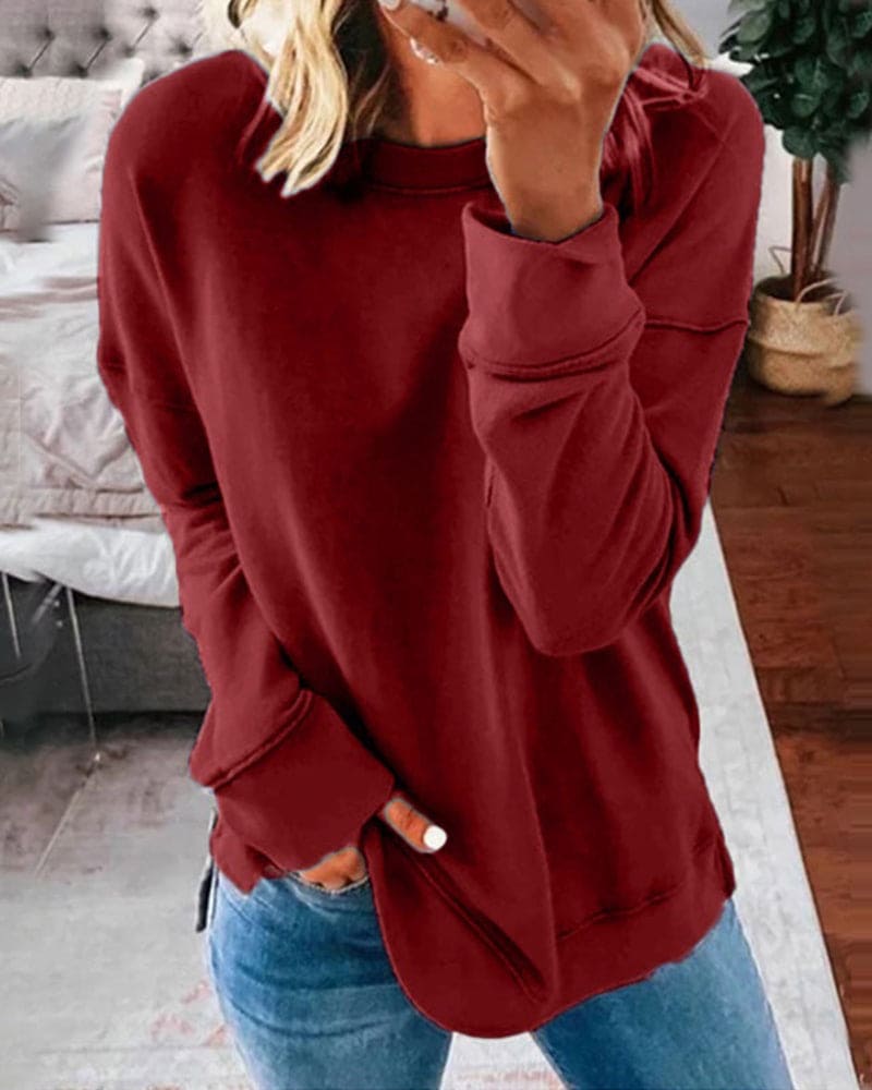 Femme portant un pull rouge bordeaux ample, manches longues, avec un jean bleu, debout dans une chambre moderne. Mode décontractée, tendance automne.