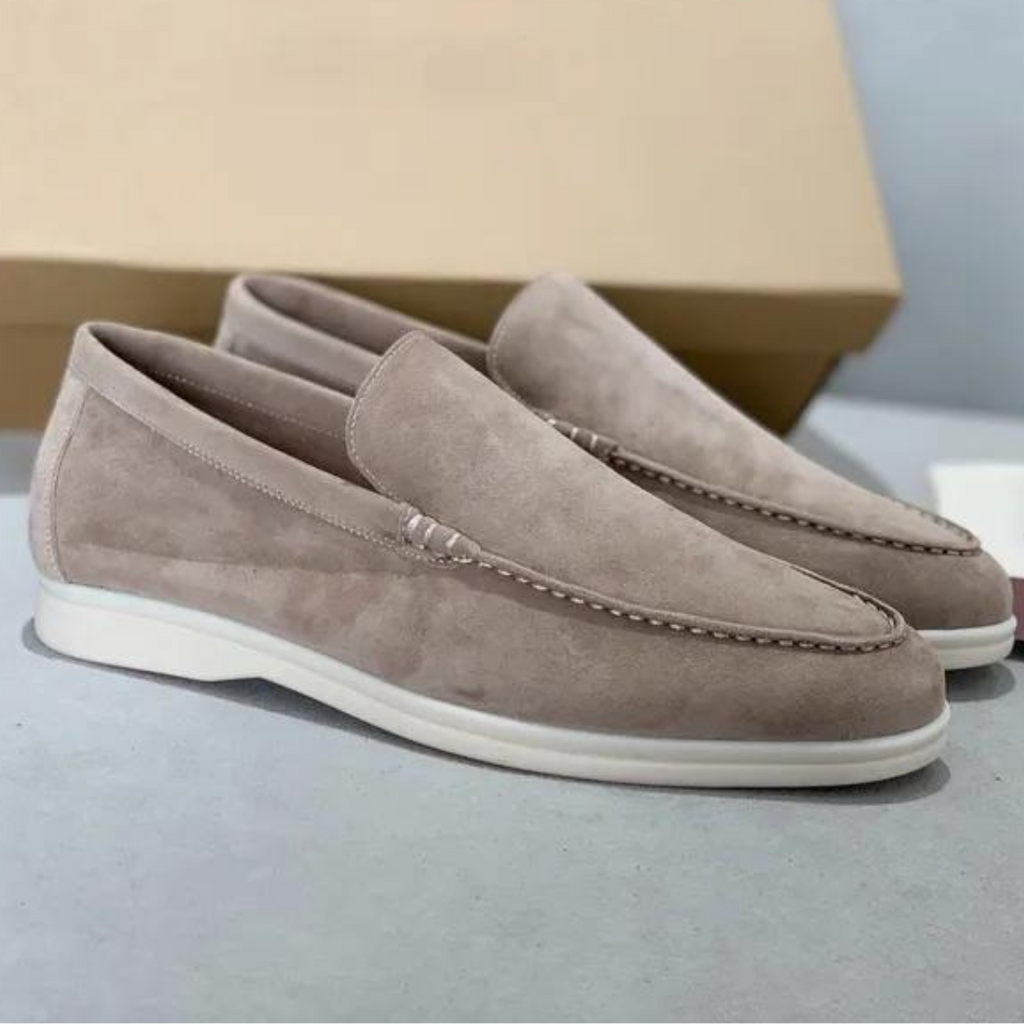 Paire de mocassins en daim beige pour homme, semelle blanche, design élégant et moderne, posée sur une surface grise avec boîte en arrière-plan.