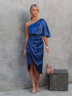 Robe bleue asymétrique élégante, femme debout contre un mur gris, sac à main beige, sandales à talons, look chic et moderne.