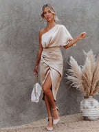 Femme en robe satinée beige asymétrique, tenant un sac blanc, posant près d'un vase avec herbes pampas, sur fond mural gris texturé.