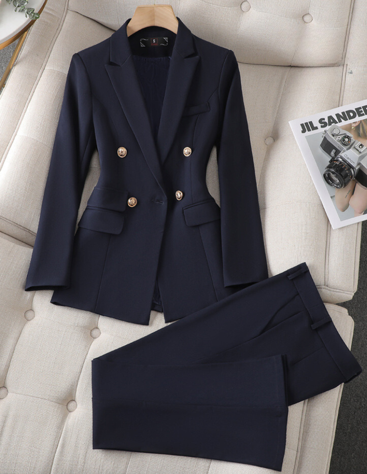 Costume femme bleu marine élégant avec boutons dorés, posé sur un canapé beige, à côté d'un magazine de photographie. Mode professionnelle chic.