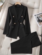 Costume noir élégant pour femme avec veste à boutons dorés et pantalon assorti, posé sur un canapé beige, à côté d'un magazine de mode.