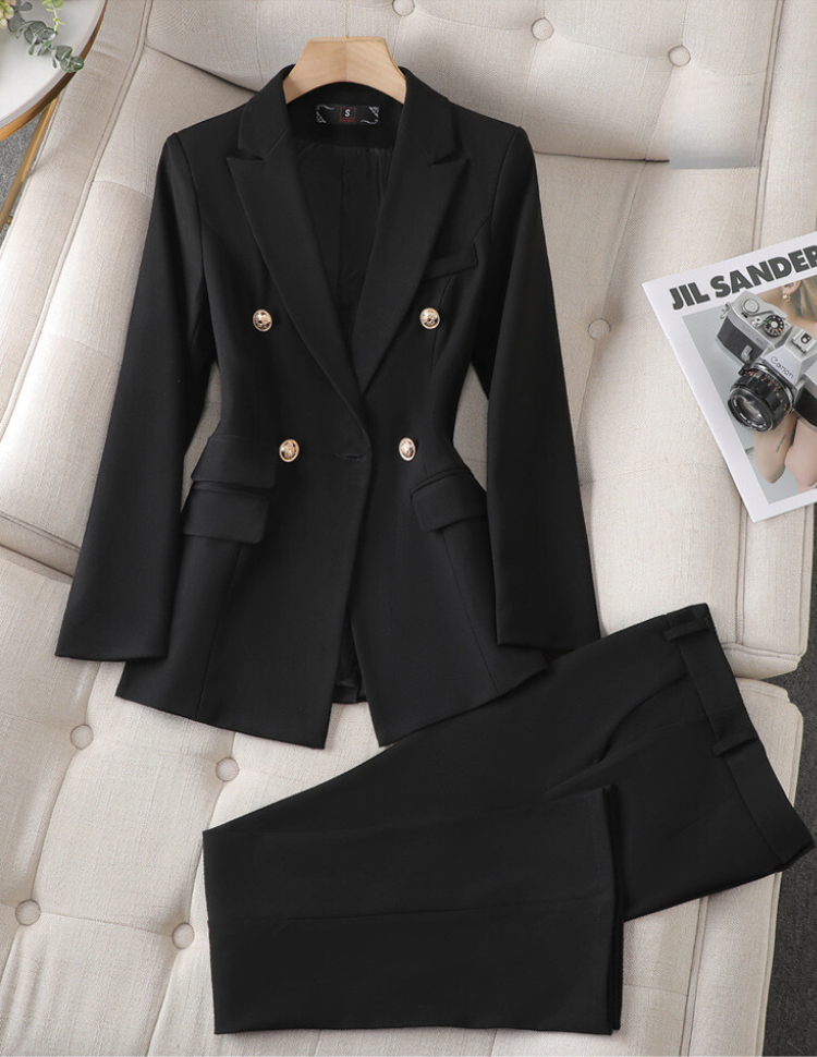 Costume noir élégant pour femme avec veste à boutons dorés et pantalon assorti, posé sur un canapé beige, à côté d'un magazine de mode.