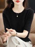 Femme portant un pull noir élégant, collier doré et boucles d'oreilles pendantes, assise sur un canapé beige. Mode féminine chic et moderne.