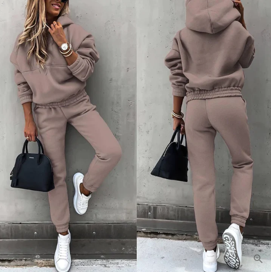 Femme en survêtement beige, baskets blanches, sac noir, posant contre un mur gris. Mode décontractée, tenue sportive élégante, style urbain.
