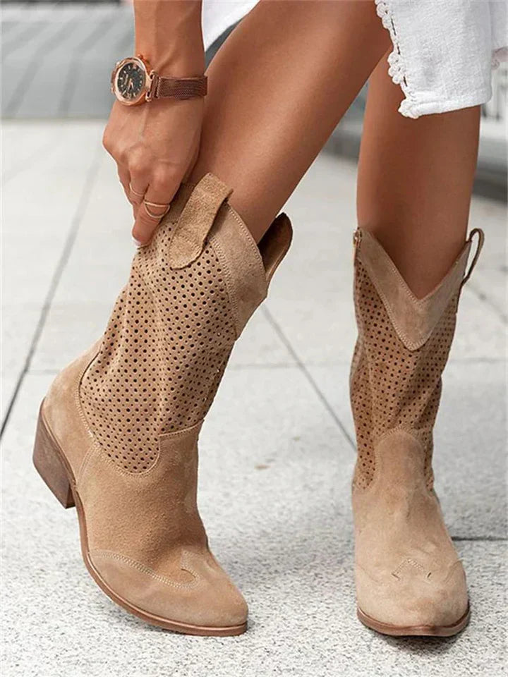 Bottes western en daim beige avec perforations, portées par une personne en robe blanche, montre-bracelet marron, sur sol pavé gris.