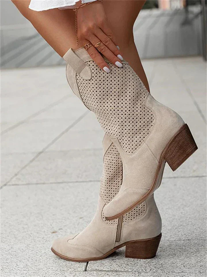 Bottes western en daim beige avec talons blocs, perforations élégantes, portées par une femme sur un trottoir urbain. Mode tendance automne.