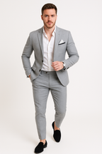Homme en costume gris élégant, chemise blanche, pochette, montre noire, chaussures mocassins noires, pose confiante, mode masculine moderne.