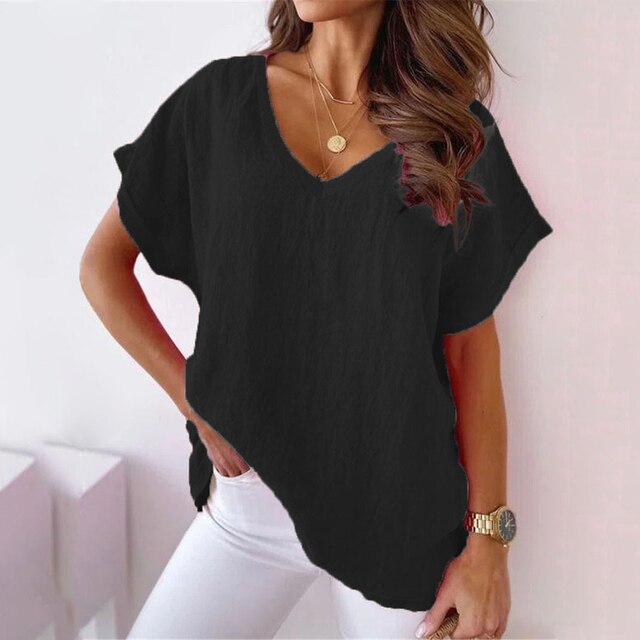 Femme portant un t-shirt noir ample avec col en V, manches courtes, accessoires dorés, posant contre un mur blanc. Mode décontractée élégante.