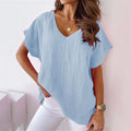 Femme portant un t-shirt bleu clair à col en V, manches courtes, avec un collier doré et une montre, posant contre un mur blanc.