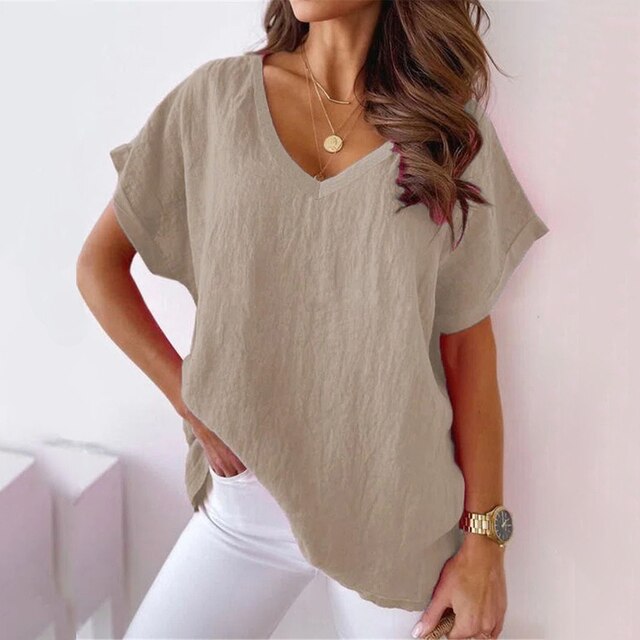 Femme portant un t-shirt beige ample en lin, col en V, manches courtes, avec un collier doré et une montre, posant contre un mur blanc.