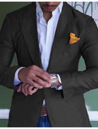 Homme en costume noir élégant, chemise blanche, mouchoir de poche orange, ajustant sa montre-bracelet argentée. Mode masculine, style professionnel.