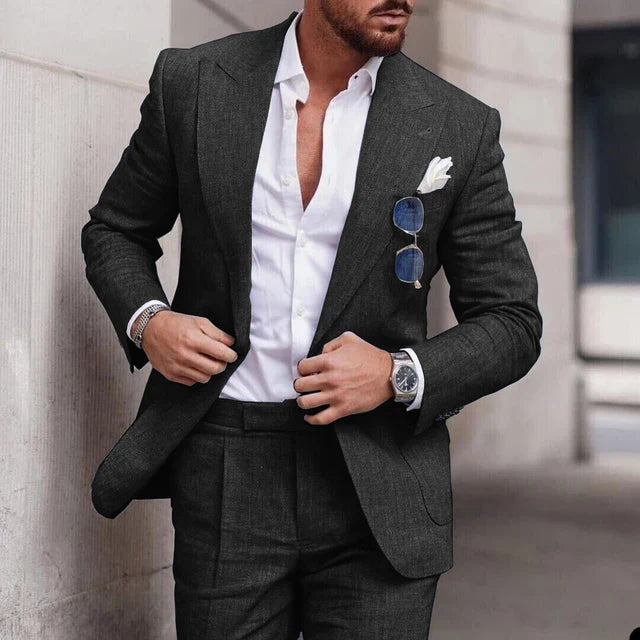 Homme en costume gris élégant, chemise blanche ouverte, lunettes de soleil accrochées, montre-bracelet argentée, style urbain chic.