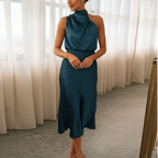 Robe élégante en satin bleu-vert, sans manches, col drapé, longueur mi-mollet, portée par une femme dans une pièce lumineuse avec rideaux blancs.