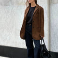 Femme portant un blazer marron en daim, t-shirt noir et jean, tenant un sac noir, posant devant un mur en marbre. Mode élégante et décontractée.