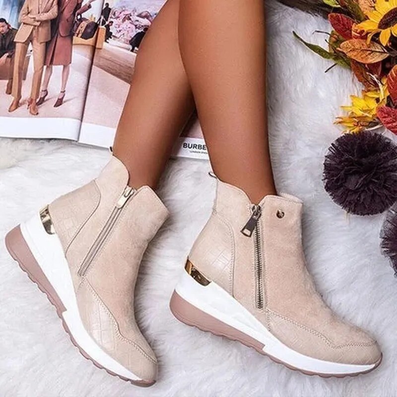 Bottes beige en daim avec fermeture éclair, semelle blanche et détails dorés, posées sur un tapis blanc, à côté d'un magazine et de fleurs d'automne.