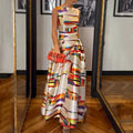 Robe longue colorée à motifs géométriques, portée par une femme tenant une pochette rouge, dans une pièce avec parquet et miroirs.