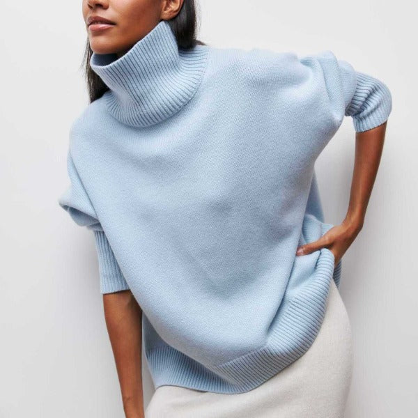 Femme portant un pull bleu ciel à col roulé, manches retroussées, sur fond blanc. Mode automne-hiver, style décontracté et élégant.