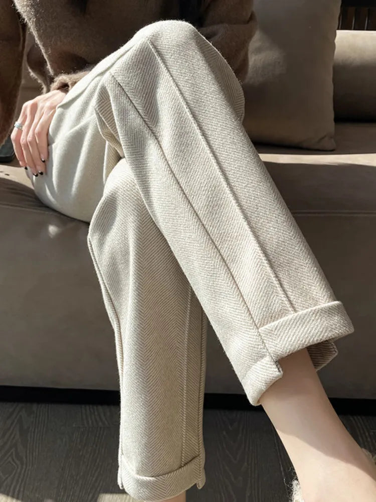 Pantalon beige en laine, coupe droite, avec revers, porté par une personne assise sur un canapé marron. Mode automne-hiver, style élégant.