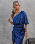 Femme en robe bleue satinée asymétrique, tenant un sac à chaîne dorée, posant devant un mur gris. Mode élégante, style chic, tendance 2023.