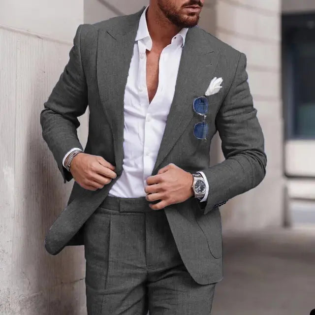 Homme élégant en costume gris, chemise blanche ouverte, lunettes de soleil bleues, montre argentée, mouchoir blanc, style mode masculine chic.