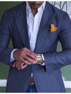 Homme élégant en costume bleu, chemise blanche, ajustant sa montre-bracelet argentée, pochette orange. Mode masculine, style chic et sophistiqué.