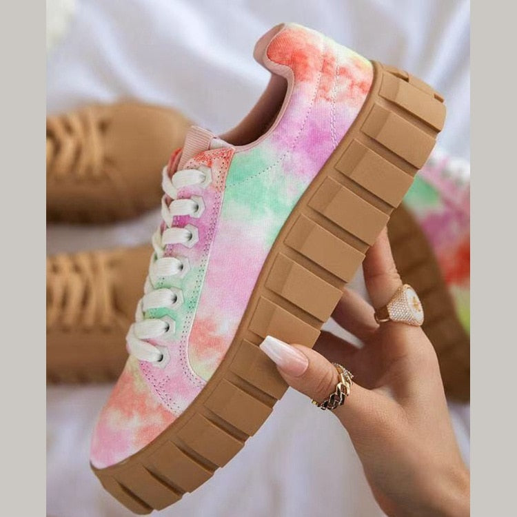 Basket colorée tie-dye pastel avec semelle épaisse marron, tenue par une main avec des ongles manucurés et bagues, sur fond flou.